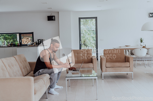 Image of African American sport man in glasses sitting at a table in a modern living room, using a laptop for business video chat, conversation with friends and entertainment