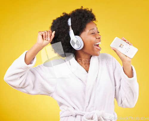 Image of Black woman, singing and happiness in a bathrobe with phone in studio isolated on a yellow background. Singer, headphones or happy female dance with mobile microphone while streaming podcast or radio