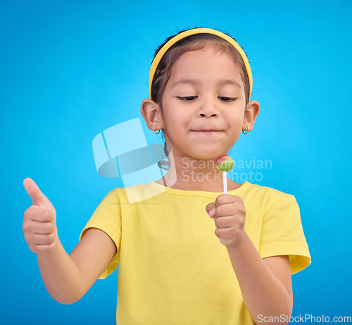 Image of Candy, happy and child with lollipop and thumbs up on blue background with happiness, smile and excited. Party, childhood and young girl with hand sign eating sweets snack, sugar and treats in studio