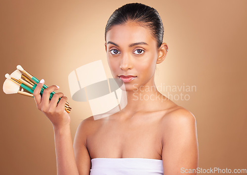 Image of Makeup, serious and brush with portrait of indian woman in studio for foundation, blush and self care. Glow, cosmetics and facial with model on brown background for shine, clean and confidence