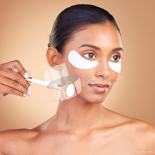 Image of Skincare, eye patch and roller with Indian woman in studio for facial, spa treatment and glow. Self care, cosmetics and hydration with female model on brown background for mask, product and youth