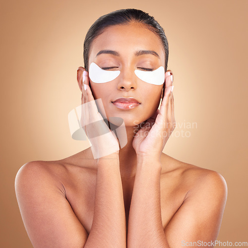 Image of Skincare, eye patch and touch with Indian woman in studio for facial, spa treatment and glow. Self care, cosmetics and hydration with female model on brown background for mask, product and youth