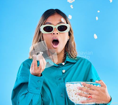 Image of 3D movie, popcorn and surprise with a woman in studio on a blue background looking shocked while eating a snack. Portrait, glasses and wow with an attractive young female watching video entertainment