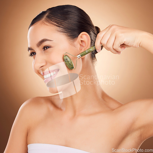 Image of Jade roller, skincare and happy woman in studio for beauty, wellness and anti aging against brown background. Facial, skin and girl relax with massaging tool, facelift and lymphatic drainage isolated