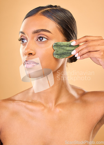 Image of Face, beauty skincare and woman with gua sha in studio isolated on a brown background. Dermatology, thinking and serious Indian female model with jade crystal or stone for healthy skin treatment.