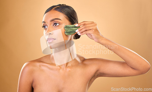 Image of Face, beauty skincare and woman with gua sha in studio isolated on a brown background. Dermatology, thinking and serious Indian female model with jade crystal or stone for healthy skin treatment.