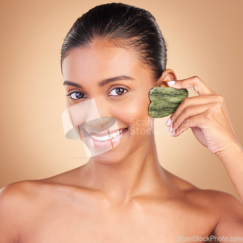 Image of Beauty, gua sha and portrait of Indian woman with smile for skincare, facial treatment and spa wellness. Salon, dermatology and girl on brown background with cosmetics, face massage and luxury stone