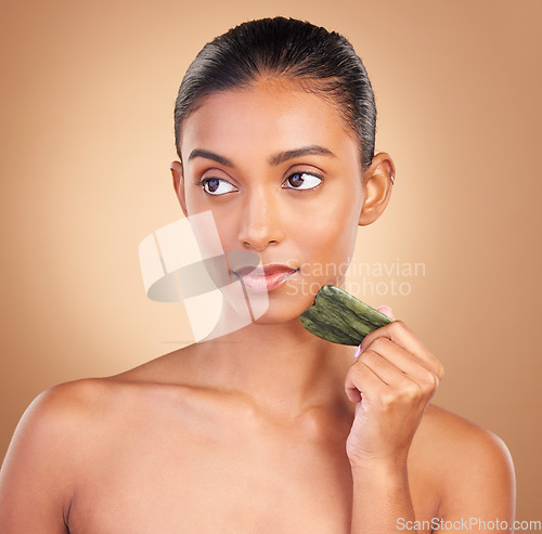 Image of Skincare, gua sha and face of Indian woman for beauty, facial treatment and wellness with spa tools. Luxury salon, dermatology and serious girl on brown background with cosmetics, massage and stone