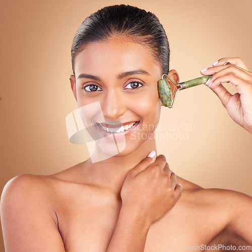 Image of Portrait, massage and skincare with a model woman in studio on a brown background for natural antiaging beauty. Face, luxury and product with an attractive young female using a roller for skin lift