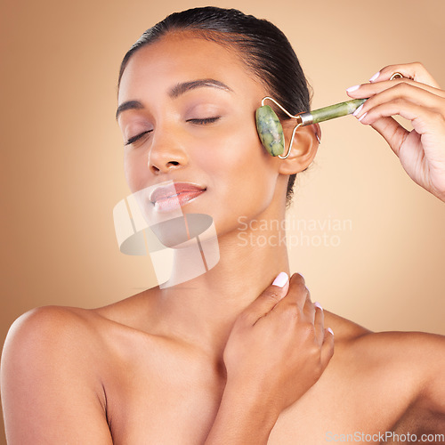 Image of Face, massage and beauty with a model woman in studio on a brown background for natural antiaging skincare. Wellness, luxury and product with an attractive young female using a roller for skin lift