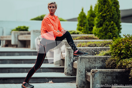 Image of Fit attractive woman in sportswear stretching one leg before jogging on the footpath outdoor in summer among greenery. Workout, sport, activity, fitness, vacation and training concept.