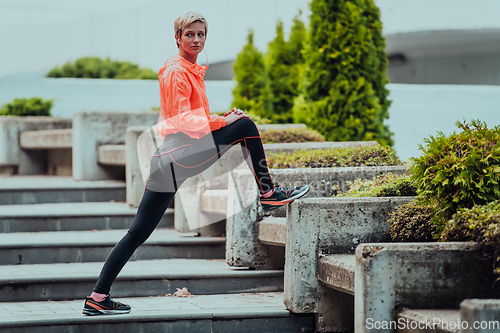Image of Fit attractive woman in sportswear stretching one leg before jogging on the footpath outdoor in summer among greenery. Workout, sport, activity, fitness, vacation and training concept.