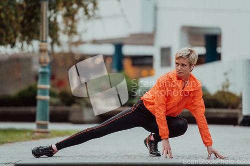 Image of Fit attractive woman in sportswear stretching one leg before jogging on the footpath outdoor in summer among greenery. Workout, sport, activity, fitness, vacation and training concept.