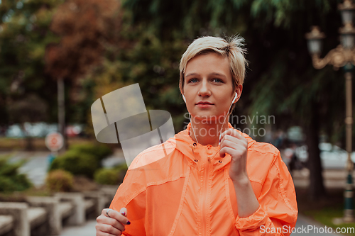 Image of a woman in a sports outfit is resting in a city environment after a hard morning workout while using noiseless headphones