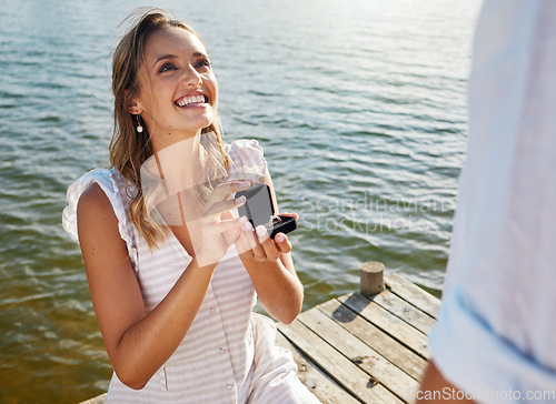 Image of Happy, love and a woman proposing to a man, getting engaged and showing a ring. Couple, excited and a young girl asking her boyfriend for marriage, kneeling to propose and ask a question by a lake