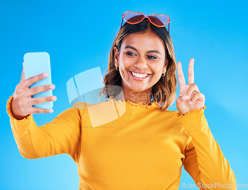 Image of Selfie smile, studio and peace sign of a woman influencer taking a profile picture for social media. Isolated, blue background and emoji hands gesture of a gen z female with feeling happy and fun