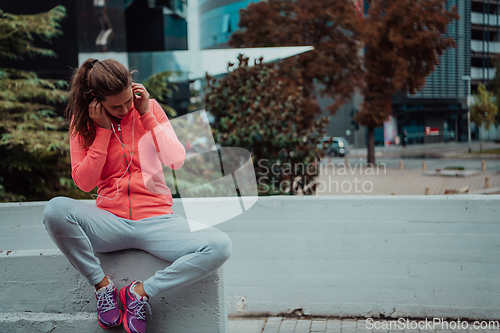 Image of a woman in a sports outfit is resting in a city environment after a hard morning workout while using noiseless headphones