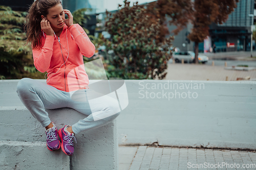 Image of a woman in a sports outfit is resting in a city environment after a hard morning workout while using noiseless headphones