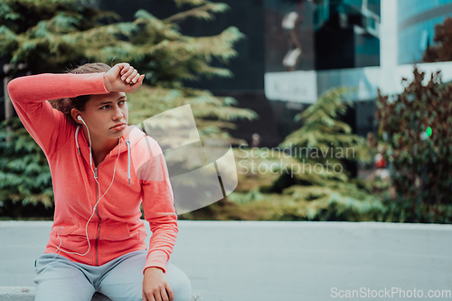 Image of a woman in a sports outfit is resting in a city environment after a hard morning workout while using noiseless headphones