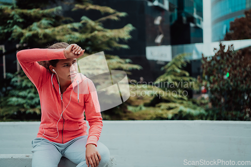 Image of a woman in a sports outfit is resting in a city environment after a hard morning workout while using noiseless headphones