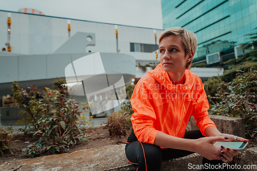 Image of A woman in sports clothes sitting in a city environment and using smartphone after a hard morning workout