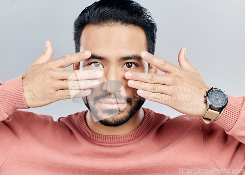 Image of Portrait, eyes and vision with a man in studio on a gray background making a masquerade hand gesture. Face, hands and focus with a handsome young male posing against a wall while touching his cheeks