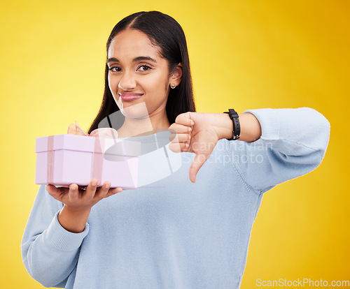 Image of Disappointed, portrait and woman with a thumbs down for a gift isolated on a yellow background. Unhappy, negative and a girl holding a present and showing a hand gesture for disappointment in studio