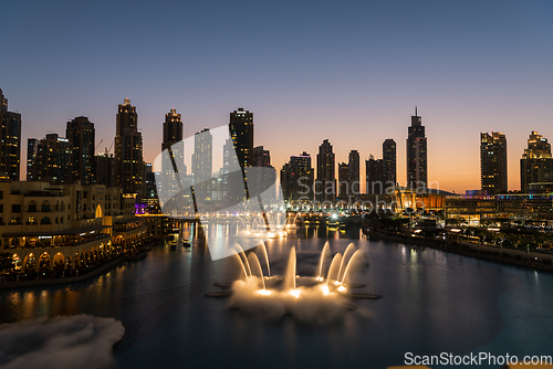 Image of Dubai singing fountains at night lake view between skyscrapers. City skyline in dusk modern architecture in UAE capital downtown.