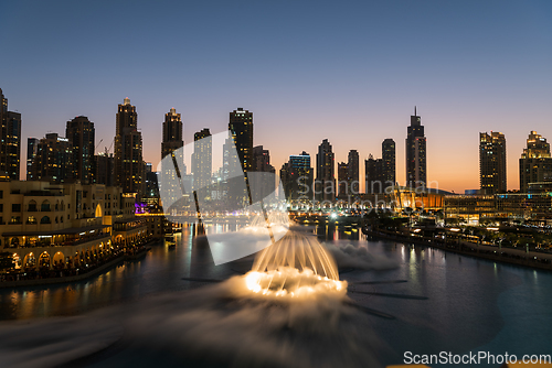 Image of Dubai singing fountains at night lake view between skyscrapers. City skyline in dusk modern architecture in UAE capital downtown.