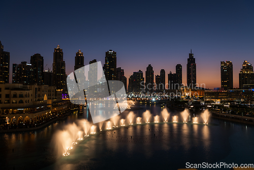 Image of Dubai singing fountains at night lake view between skyscrapers. City skyline in dusk modern architecture in UAE capital downtown.