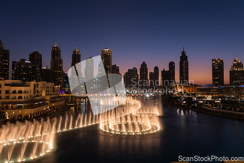 Image of Dubai singing fountains at night lake view between skyscrapers. City skyline in dusk modern architecture in UAE capital downtown.