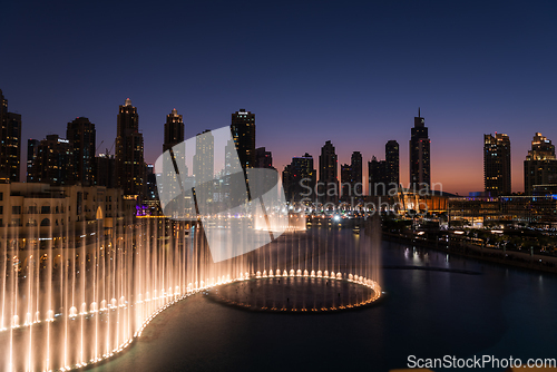 Image of Dubai singing fountains at night lake view between skyscrapers. City skyline in dusk modern architecture in UAE capital downtown.