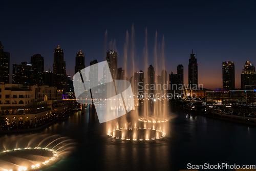 Image of Dubai singing fountains at night lake view between skyscrapers. City skyline in dusk modern architecture in UAE capital downtown.