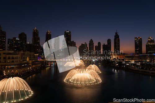 Image of Dubai singing fountains at night lake view between skyscrapers. City skyline in dusk modern architecture in UAE capital downtown.