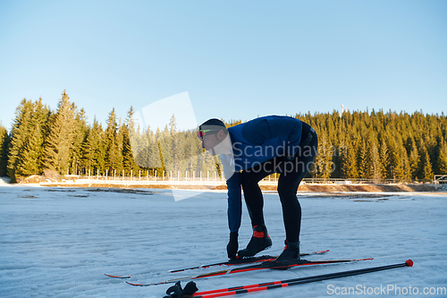 Image of Handsome male athlete with cross country skis preparing equipment for training in a snowy forest. Checking smartwatch. Healthy winter lifestyle.