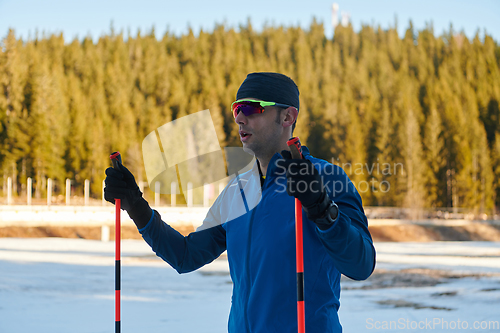 Image of Handsome male athlete with cross country skis preparing equipment for training in a snowy forest. Checking smartwatch. Healthy winter lifestyle.