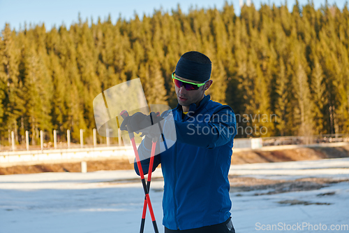 Image of Handsome male athlete with cross country skis, taking fresh breath and having break after hard workout training in a snowy forest. Checking smartwatch. Healthy winter lifestyle