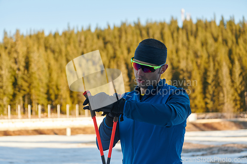 Image of Handsome male athlete with cross country skis, taking fresh breath and having break after hard workout training in a snowy forest. Checking smartwatch. Healthy winter lifestyle