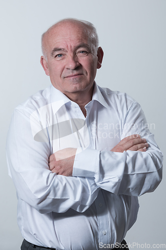 Image of Confident senior man in white shirt crossing hands on chest and looking at camera while standing against gray background. Self confident senior isolated white studio shoot.