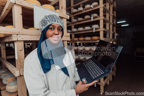 Image of African American Muslim businesswoman checking product quality and entering data into a laptop at a local cheese manufacturing company