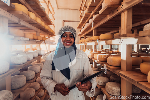 Image of African American Muslim businesswoman checking product quality and entering data into a laptop at a local cheese manufacturing company