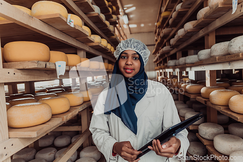 Image of African American Muslim businesswoman checking product quality and entering data into a laptop at a local cheese manufacturing company