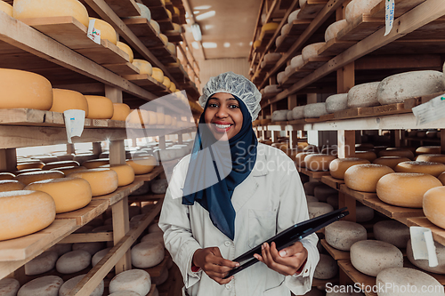 Image of African American Muslim businesswoman checking product quality and entering data into a laptop at a local cheese manufacturing company