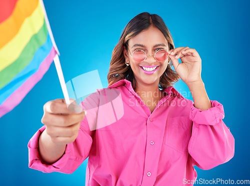 Image of Woman, lgbtq and pride flag in studio portrait, sunglasses or icon with smile for human rights by blue background. Gen z girl, young lesbian student and wave sign for solidarity, freedom or inclusion
