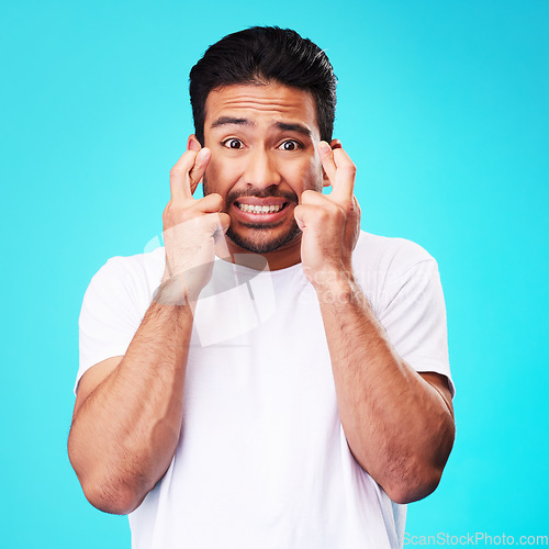 Image of Fear, hope and portrait of man fingers crossed, anxiety or nervous due to crisis or stress isolated in studio blue background. Worry, suspense and young male person hands in face for mistake reaction