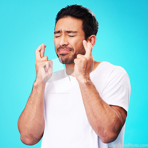 Image of Praying, hope and man fingers crossed, anxiety or nervous due to crisis or stress isolated in studio blue background. Fear, suspense and young male person hands in face for mistake or risk reaction