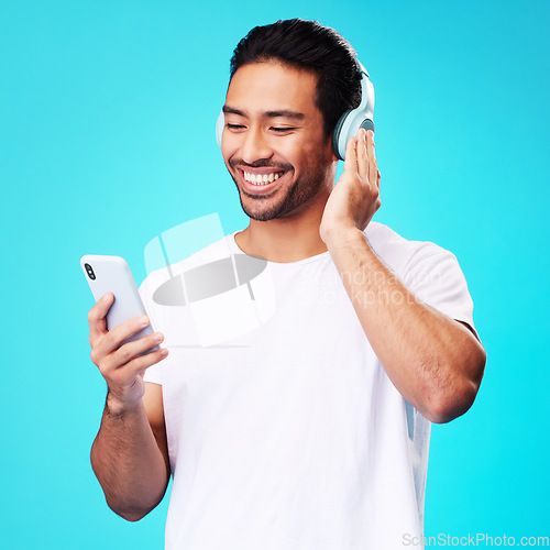 Image of Headphones, music and Asian man with smartphone for listening in studio isolated on a blue background. Radio, smile and person with mobile, sound or hearing podcast, audio or social media on internet