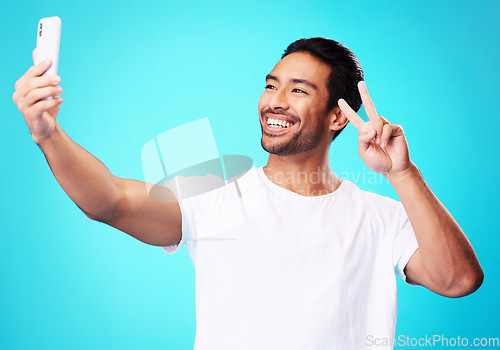 Image of Happy, peace sign and a man with a phone on a blue background for communication or social media. Smile, contact and a male influencer with an emoji hand and a mobile while live streaming in studio