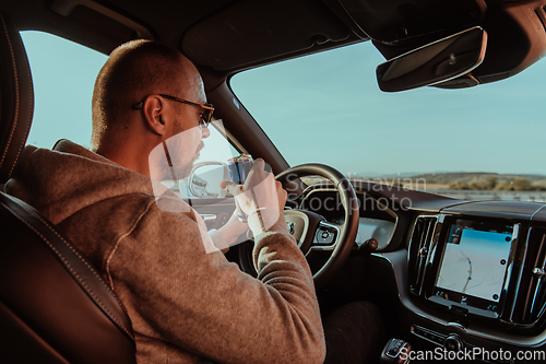 Image of A tired man drinking acoffee while driving a car at sunset. Tired travel and long drive