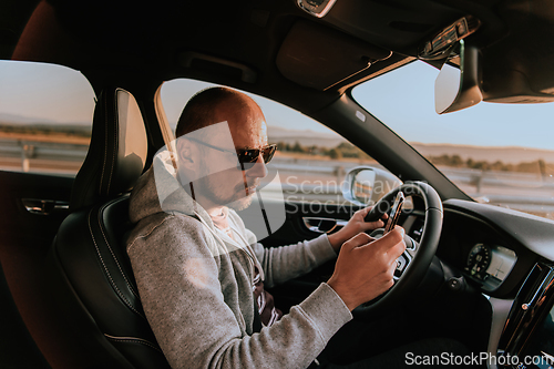 Image of A man with a sunglasses driving a car and type a message on smartphone at sunset. The concept of car travel